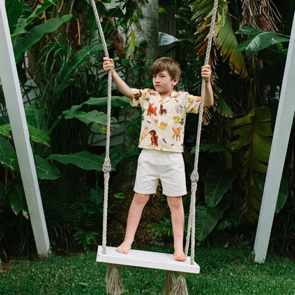 A young boy stands barefoot on a white rope swing in a lush garden, wearing a cream short-sleeve t-shirt with a colorful dog print and light beige shorts.