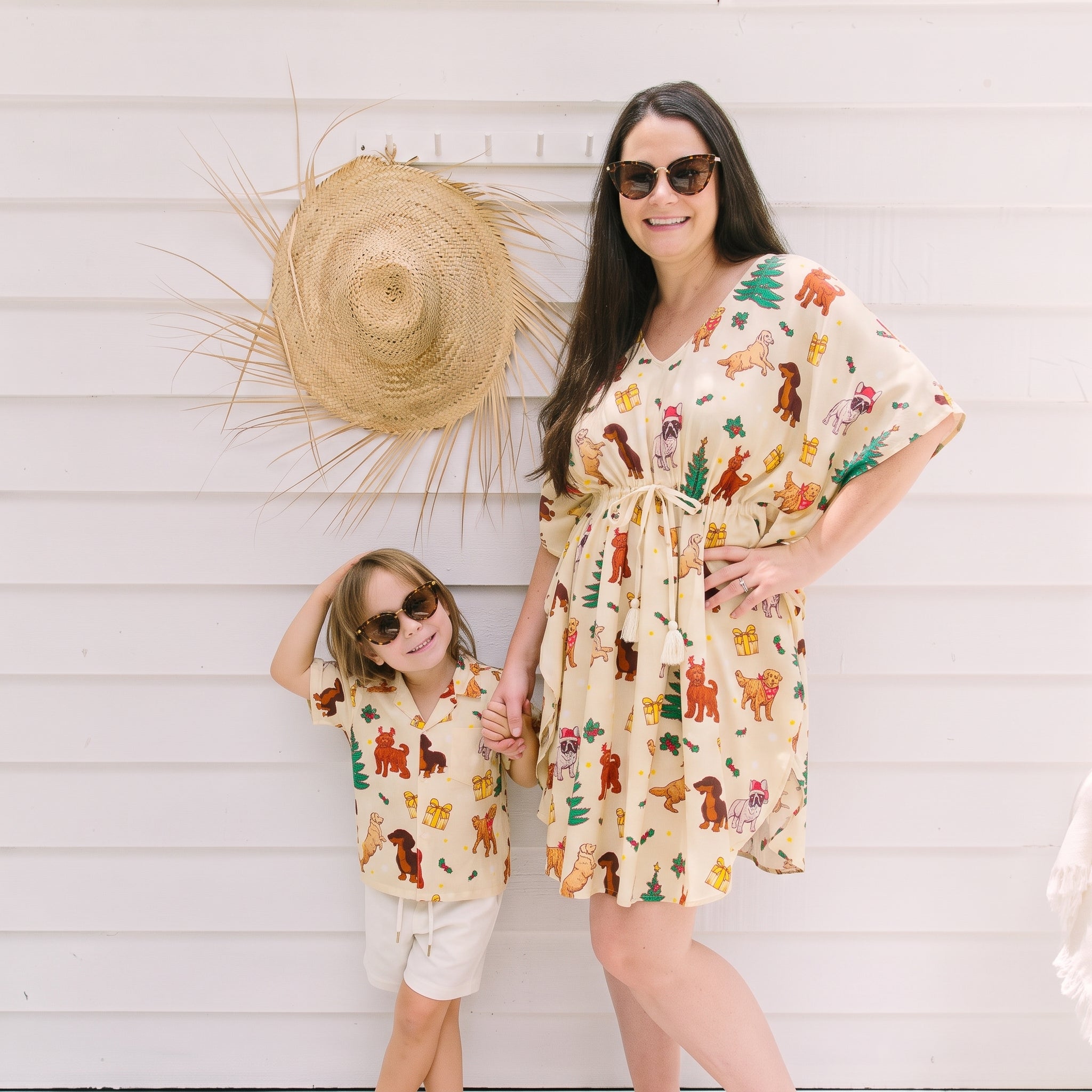 A woman and a child wearing matching cream-colored short-sleeve dresses with a colorful woodland animal print, standing against a white siding wall with a straw hat hanging on the wall.