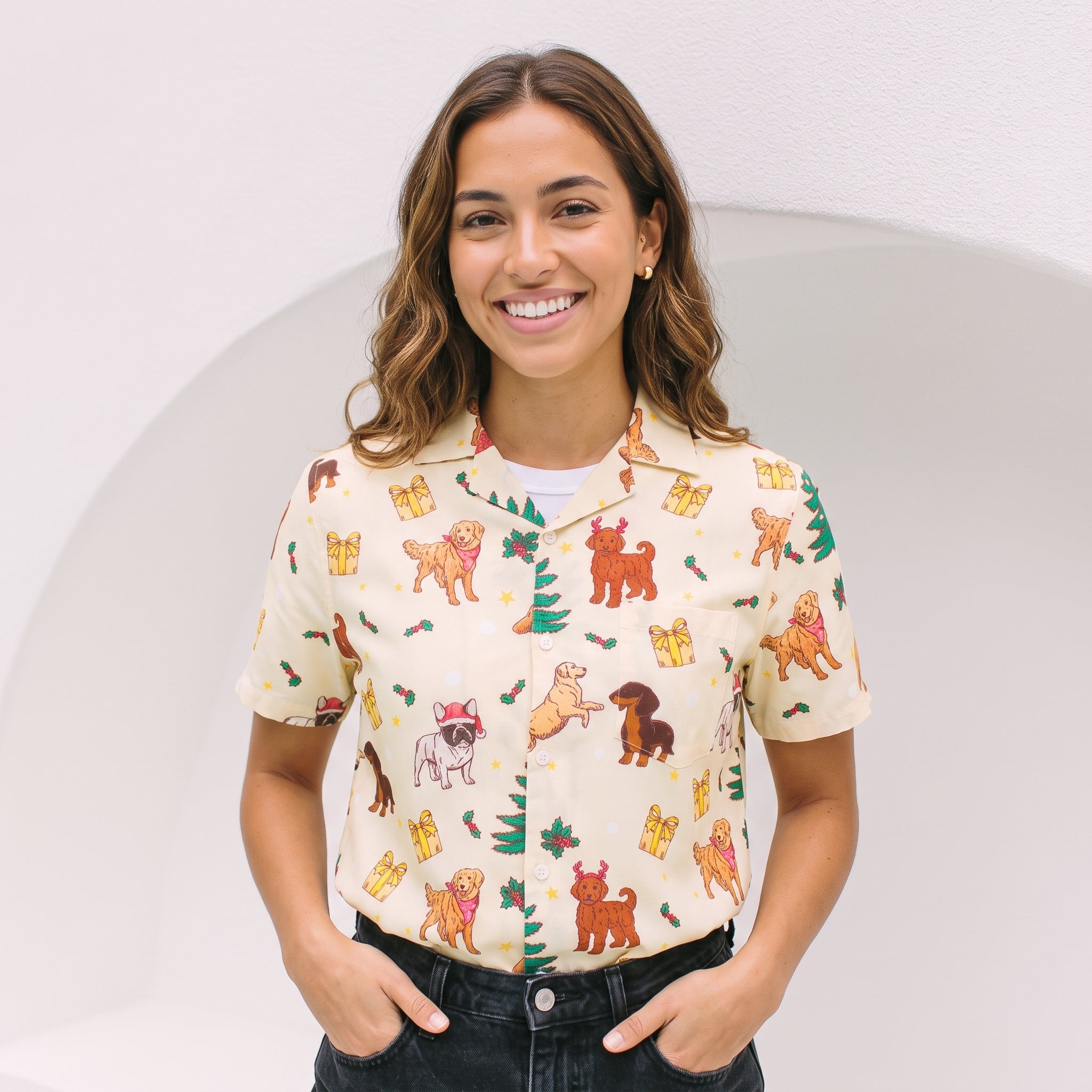 A smiling woman wearing a short-sleeve button-up shirt with a festive Christmas dog print, paired with dark jeans, standing against a clean white background with a subtle arch.