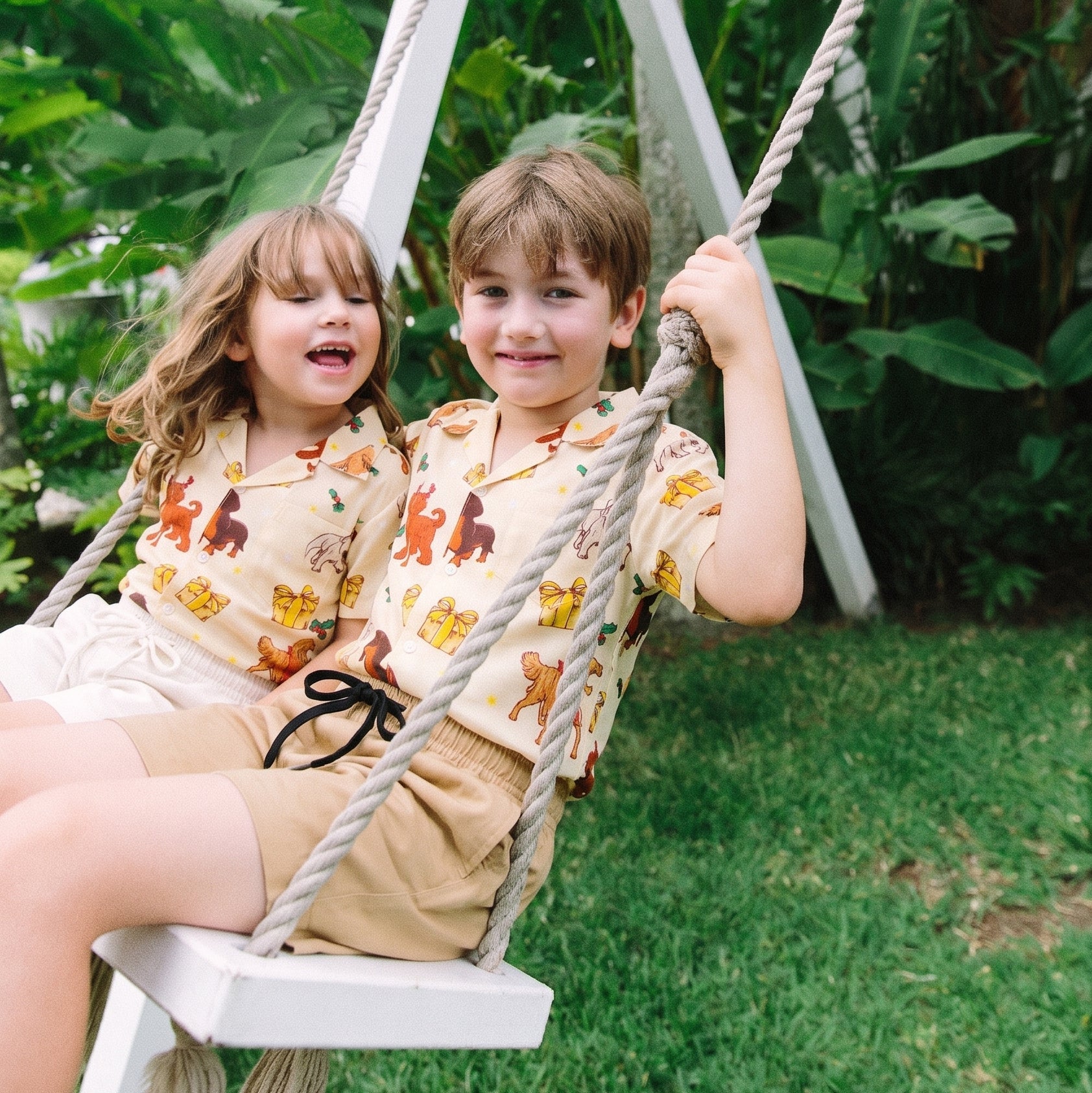 Two children sitting together on a white rope swing in a garden, wearing matching beige short-sleeve button-up shirts with a colorful cartoon animal print and tan shorts.