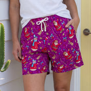 A woman stands outdoors in front of a white house wall and a yellow door, wearing bright purple patterned shorts with a white tank top. The shorts have a colorful holiday print with Santa Claus, reindeer, snowflakes, and other festive motifs.