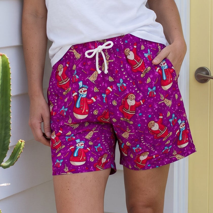 A woman stands outdoors in front of a white house wall and a yellow door, wearing bright purple patterned shorts with a white tank top. The shorts have a colorful holiday print with Santa Claus, reindeer, snowflakes, and other festive motifs.