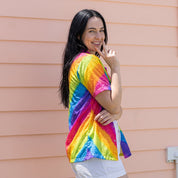 A woman with long black hair stands in front of a peach-colored horizontal siding wall, wearing a short-sleeve rainbow tie-dye shirt and white shorts.