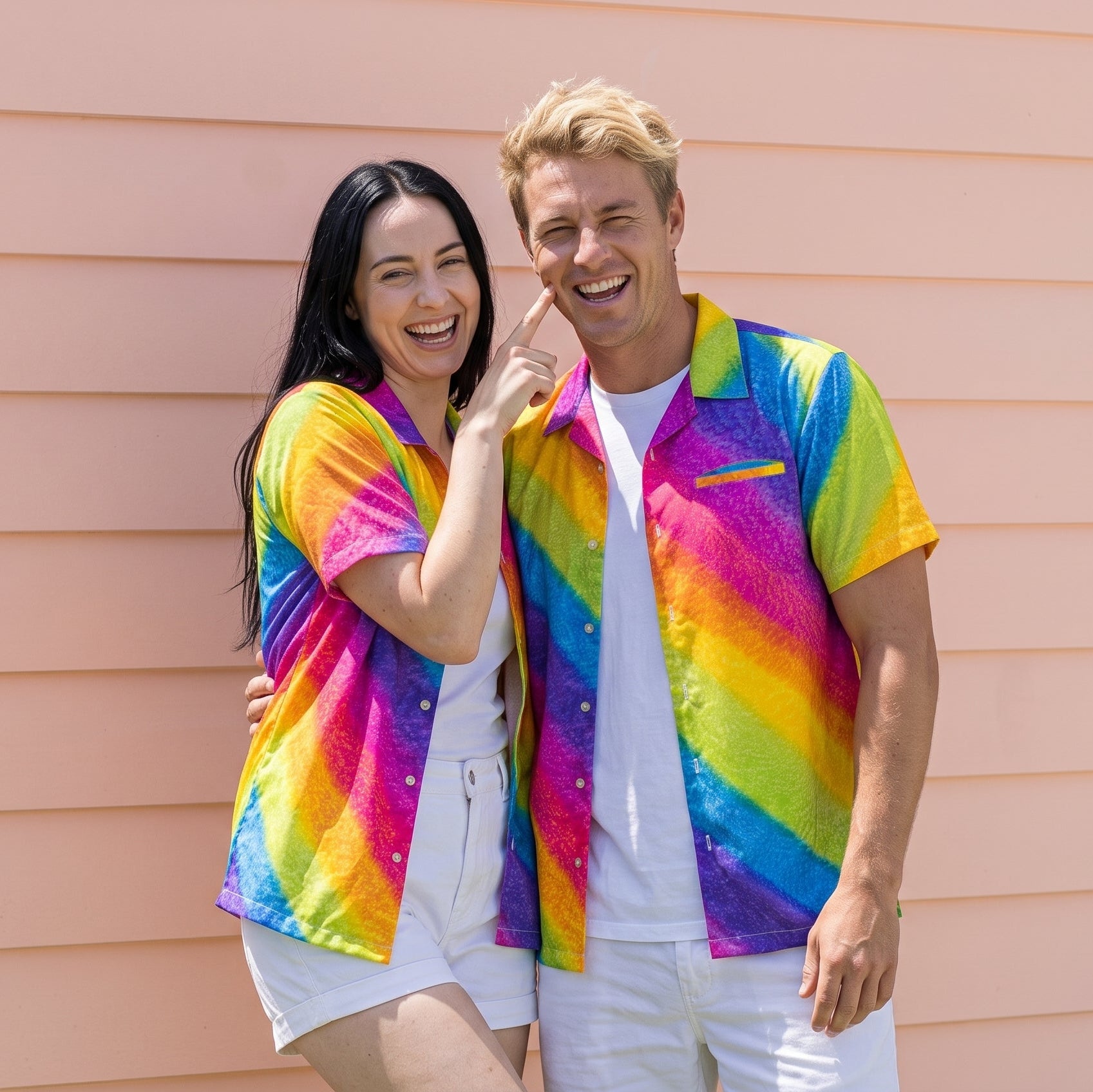 A man and a woman standing side-by-side in front of a peach-colored house wall, wearing matching short-sleeve button-up shirts with a vibrant rainbow tie-dye pattern.