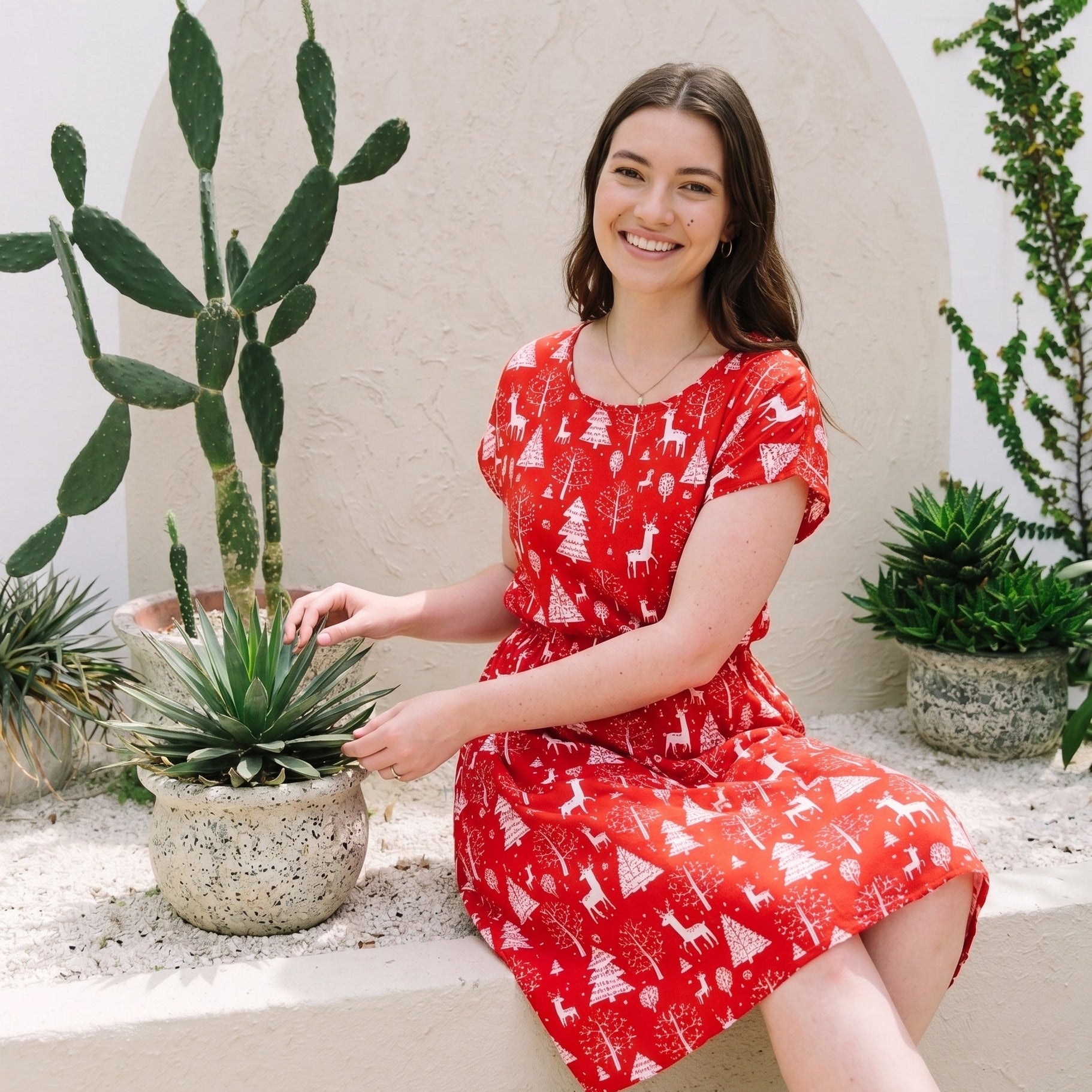 A woman wearing a red short-sleeve dress with a white Christmas tree pattern sits on a beige stone ledge next to potted cacti and succulents against a textured beige arch wall.