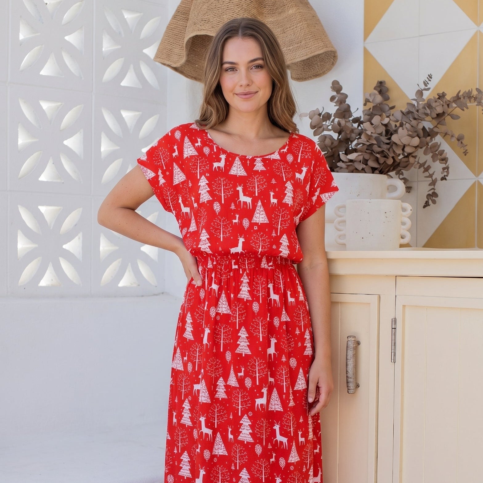 A woman modeling a red short-sleeve maxi dress with a white Christmas tree pattern, standing in a brightly lit room with white and yellow geometric wall panels.