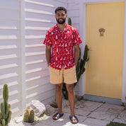 A man stands outdoors in front of a white house wall and yellow door, wearing a red short-sleeve button-up shirt with a white tropical tree print, tan shorts, and black slide sandals.