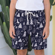 A young boy stands outdoors in front of a white house wall and a pale yellow door, wearing a plain white short-sleeve T-shirt and navy shorts with a white tree print and drawstring.