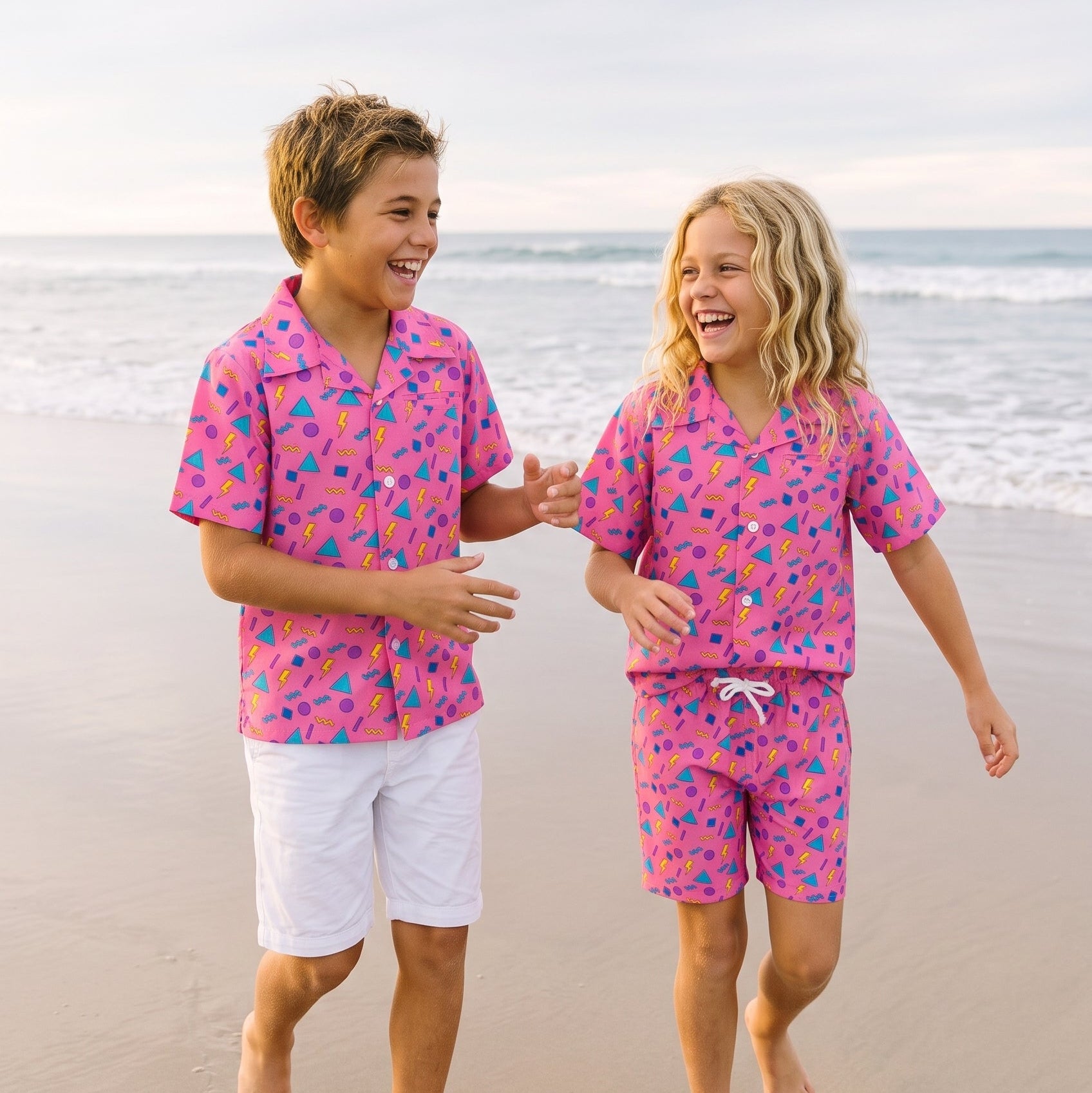 Two children walking hand in hand on a sandy beach wearing matching pink short-sleeve camp shirts and shorts sets with a colorful abstract pattern.