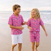 Two children walking hand in hand on a sandy beach wearing matching pink short-sleeve camp shirts and shorts sets with a colorful abstract pattern.