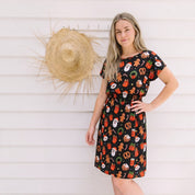 A woman modeling a short-sleeve black dress with a colorful Christmas-themed print of Santa Claus, reindeer, and ornaments, standing against a white wood-paneled wall with a straw hat hanging on the wall.