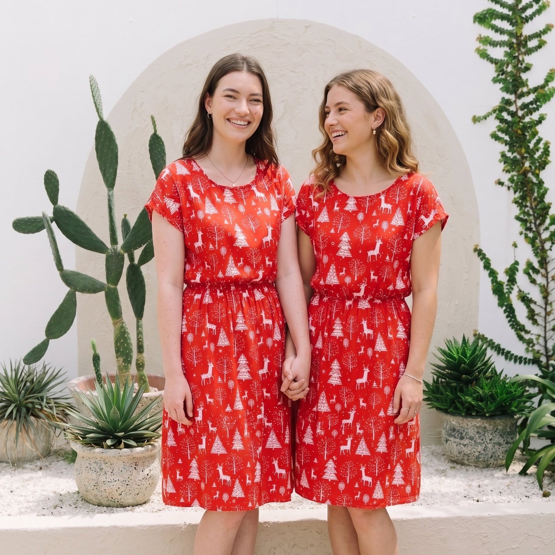 Two women standing side-by-side in matching red short-sleeve dresses with a white Christmas tree and reindeer print, posing in front of a white wall with potted plants.