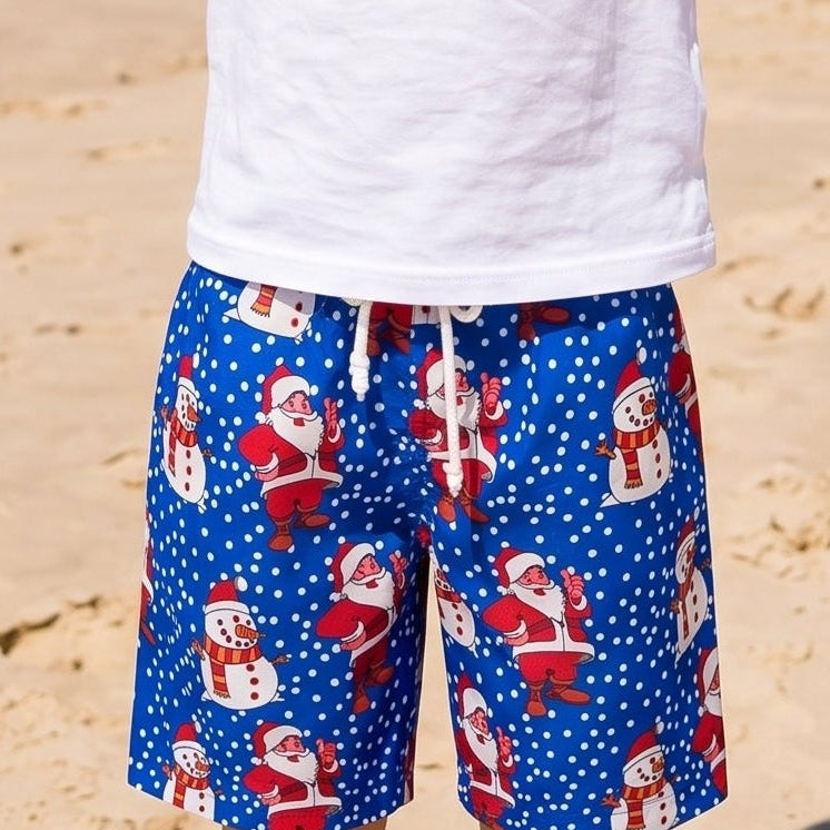 A young boy stands on a sandy beach with the ocean and blue sky behind him, balancing a pineapple on his head. He is wearing a plain white t-shirt and blue patterned shorts with Santa Claus designs.