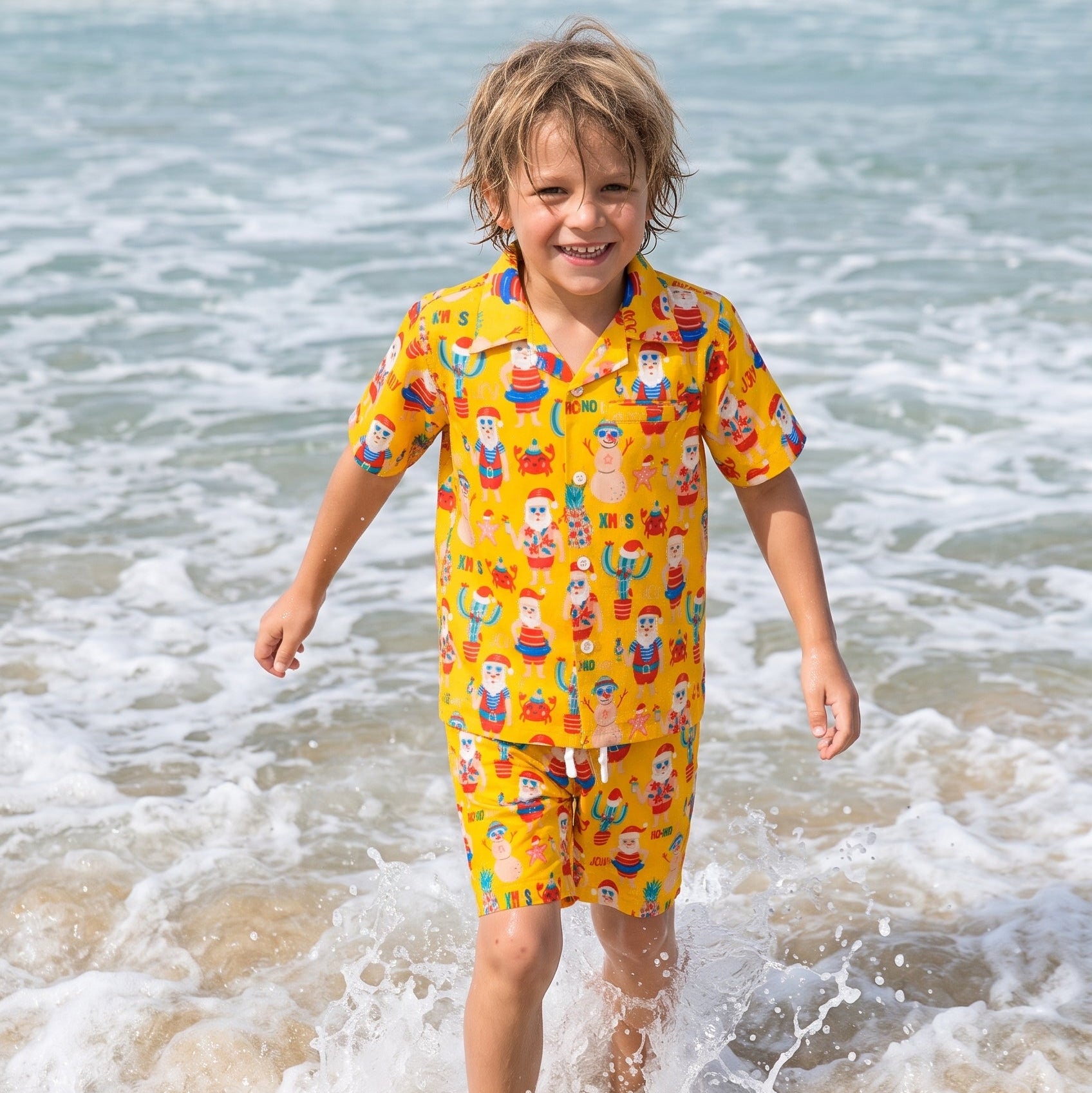 A young boy with blonde hair stands barefoot in the shallow surf of a beach, wearing a bright yellow two-piece pajama set with a colorful cartoon print.