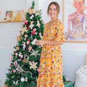 A woman wearing a bright yellow midi dress with a colorful Christmas print stands next to a decorated Christmas tree in a bright room with holiday gifts underneath.