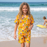 A young girl walking on a sandy beach wearing a yellow two-piece pajama set with a colorful cartoon print. The set includes a short-sleeve button-up shirt and matching shorts.
