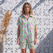 A woman stands outdoors in front of a white geometric wall, wearing a matching short-sleeve button-up shirt and shorts set with a colorful tropical floral print. She is wearing white sneakers, with potted dried grass on the left side of the frame.