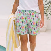 A woman walking beside a pool wearing a white t-shirt and colorful tropical print swim shorts, holding a yellow and white striped towel.