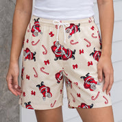 A woman modeling a white ribbed tank top and beige holiday-themed shorts with a Santa Claus and candy cane print, standing on a white porch.