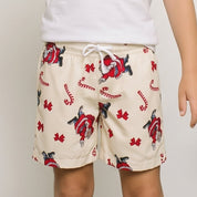 A young boy stands barefoot in a studio setting, wearing a plain white t-shirt and beige swim trunks with a red and blue flamingo pattern. He is posing in front of a white wall with two large potted green plants on either side.