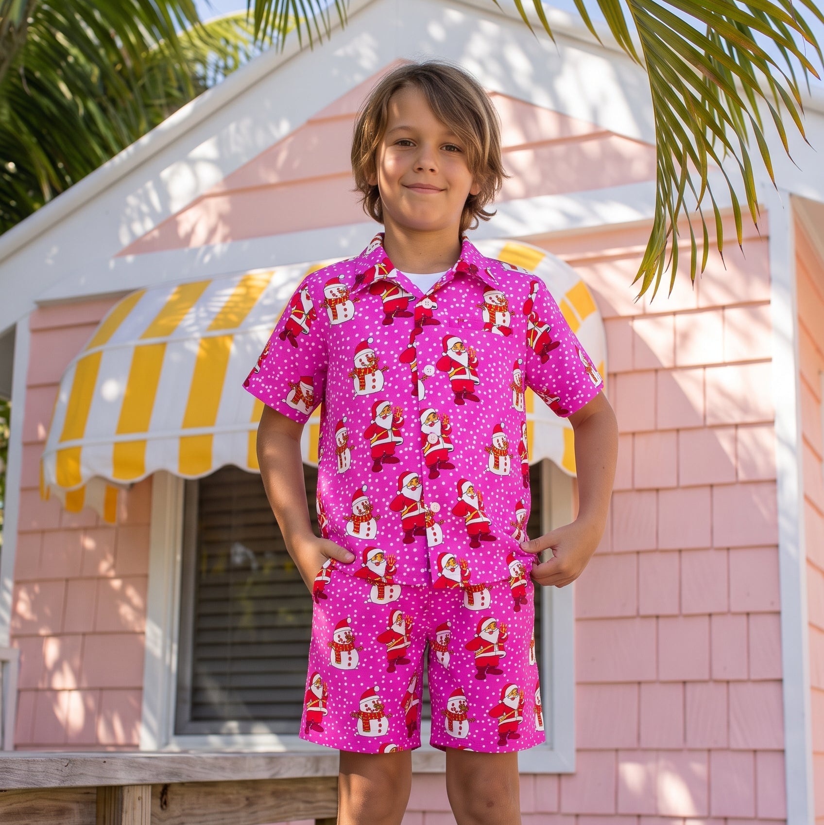 A young boy stands on a wooden deck in front of a pink house with a yellow-and-white awning, wearing a bright pink short-sleeve button-up shirt and matching shorts with a red-and-white Santa Claus gift print.