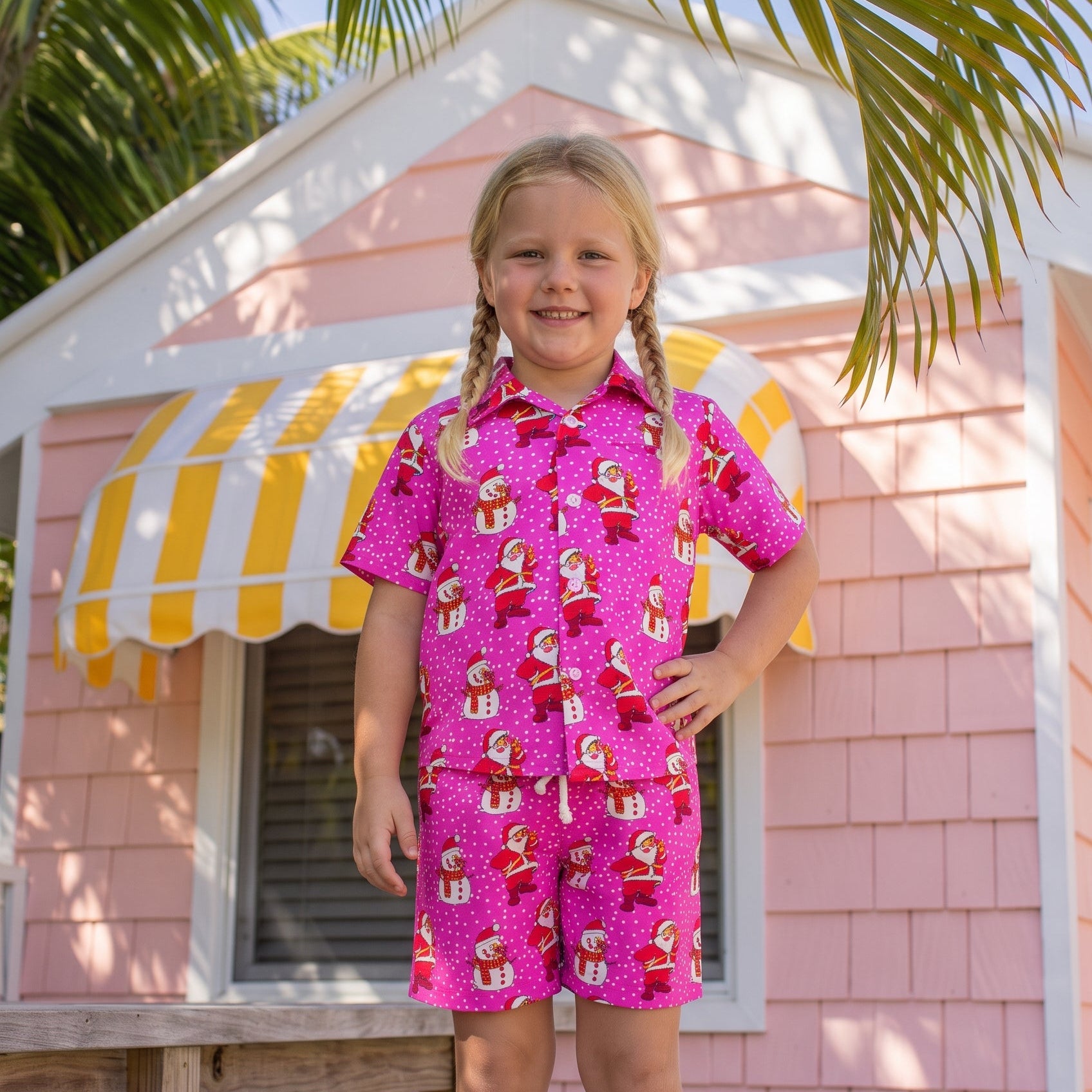 A young girl with blonde braids stands on a wooden deck in front of a pink house with a yellow and white striped awning, wearing a bright pink short-sleeve collared shirt and matching shorts set with a Santa Claus gift pattern.