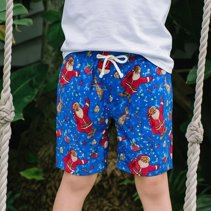 A young boy stands barefoot on a white rope swing in a lush garden setting. He is wearing a plain white t-shirt and blue swim trunks with a red and white flamingo pattern.