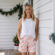 A woman stands on a porch wearing a white sleeveless top and pink-and-white patterned shorts, with a white house, wooden deck, and holiday garland in the background.
