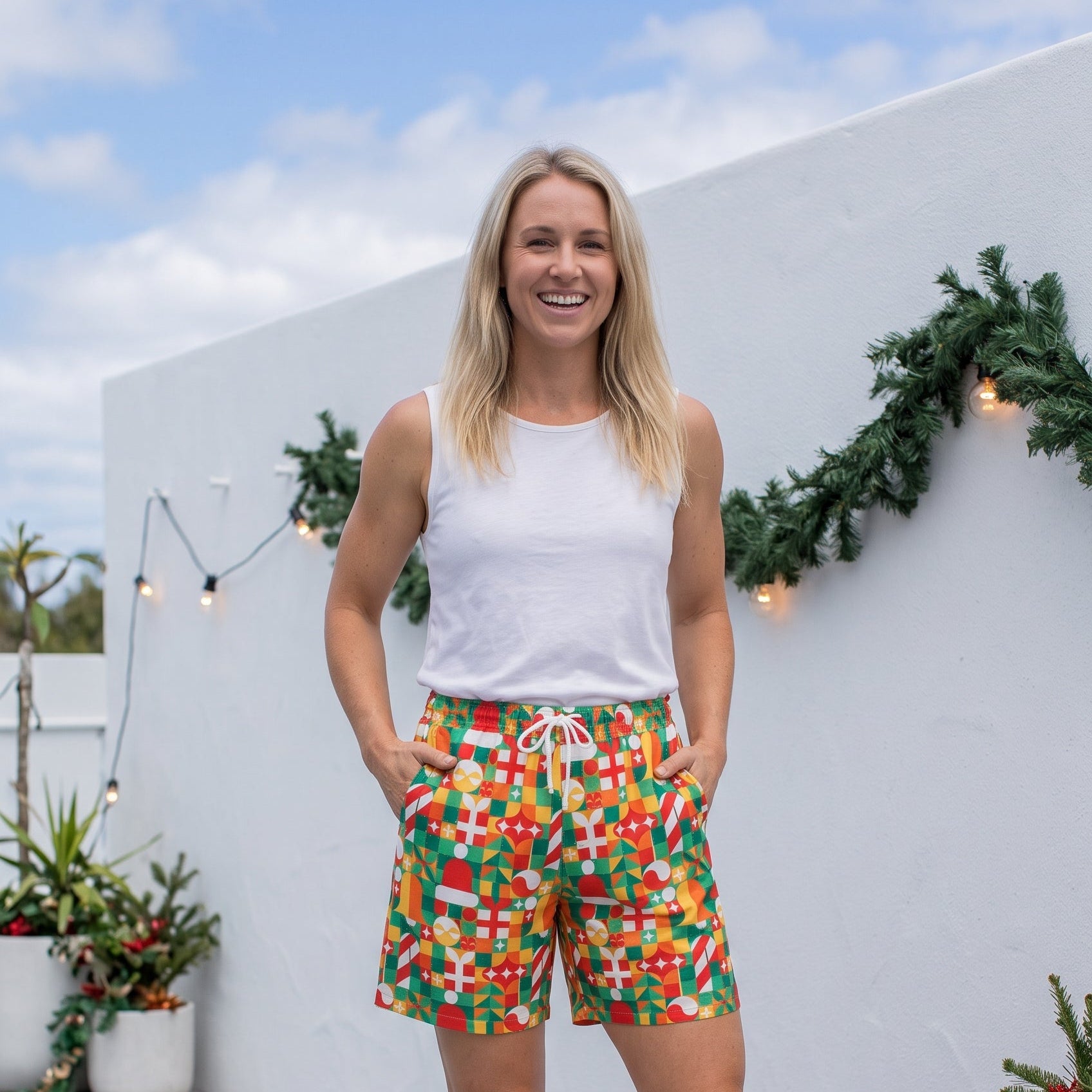 A woman stands on a light gray wooden deck wearing colorful patterned shorts, a white tank top, and black slide sandals, posing in front of a white wall decorated with green garlands.