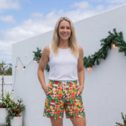 A woman stands on a light gray wooden deck wearing colorful patterned shorts, a white tank top, and black slide sandals, posing in front of a white wall decorated with green garlands.