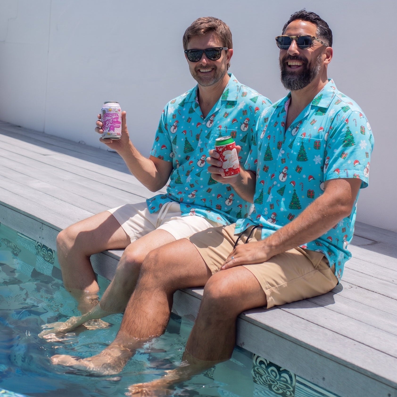 Two men sitting on the edge of a swimming pool wearing matching turquoise Hawaiian shirts with a red and yellow tropical pattern, khaki shorts, and sunglasses, holding drinks.
