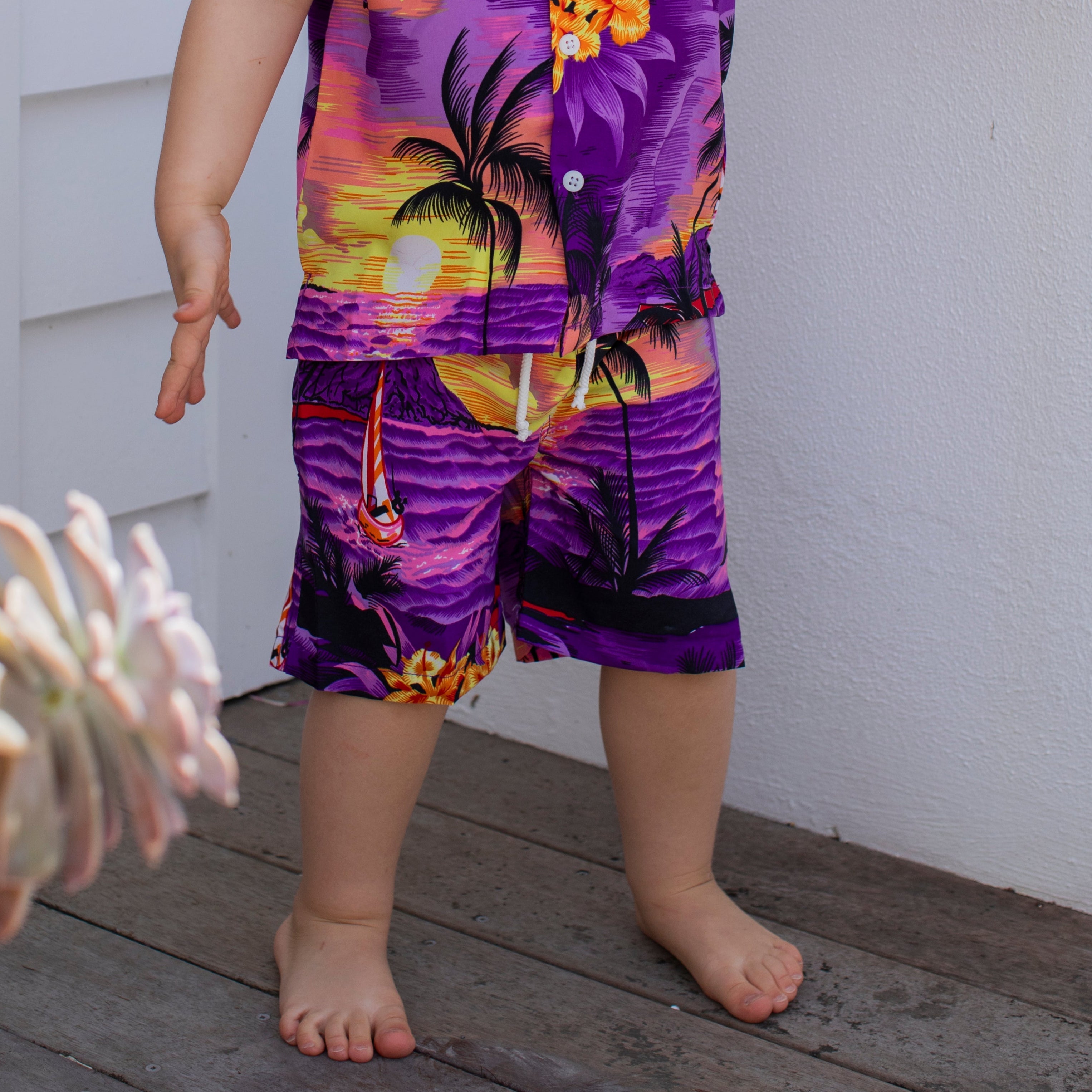 A barefoot child stands on a wooden floor, wearing Sunset Purple Kids Shorts paired with a vibrant tropical outfit featuring palm trees and sunset designs—ideal for festive family gatherings. Only the lower half of the child's body is visible.