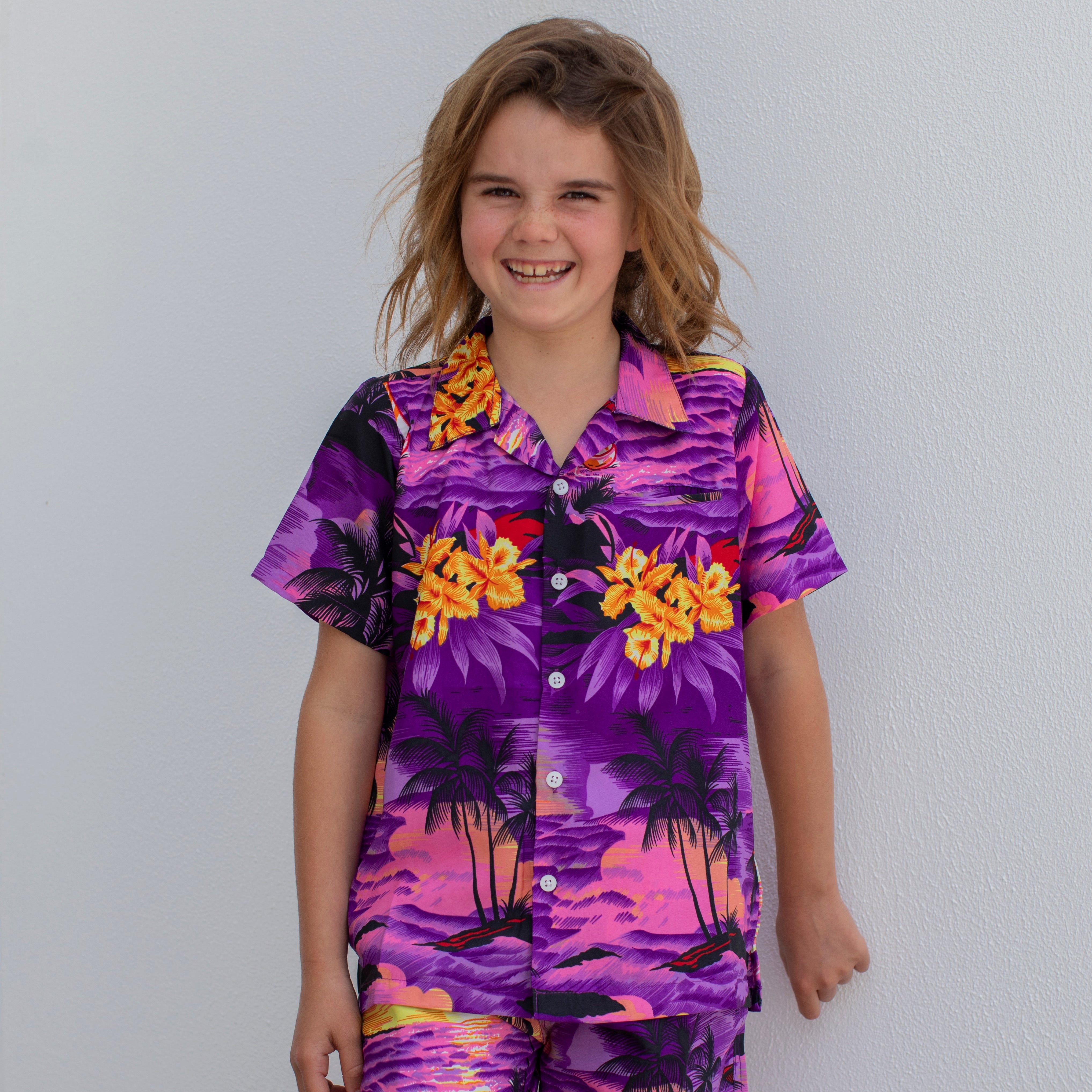 A smiling child with tousled hair wears the Sunset Purple Kids Hawaiian Shirt and Shorts Set, featuring a tropical print in purple, pink, and yellow—the perfect summer outfit against a plain white wall.