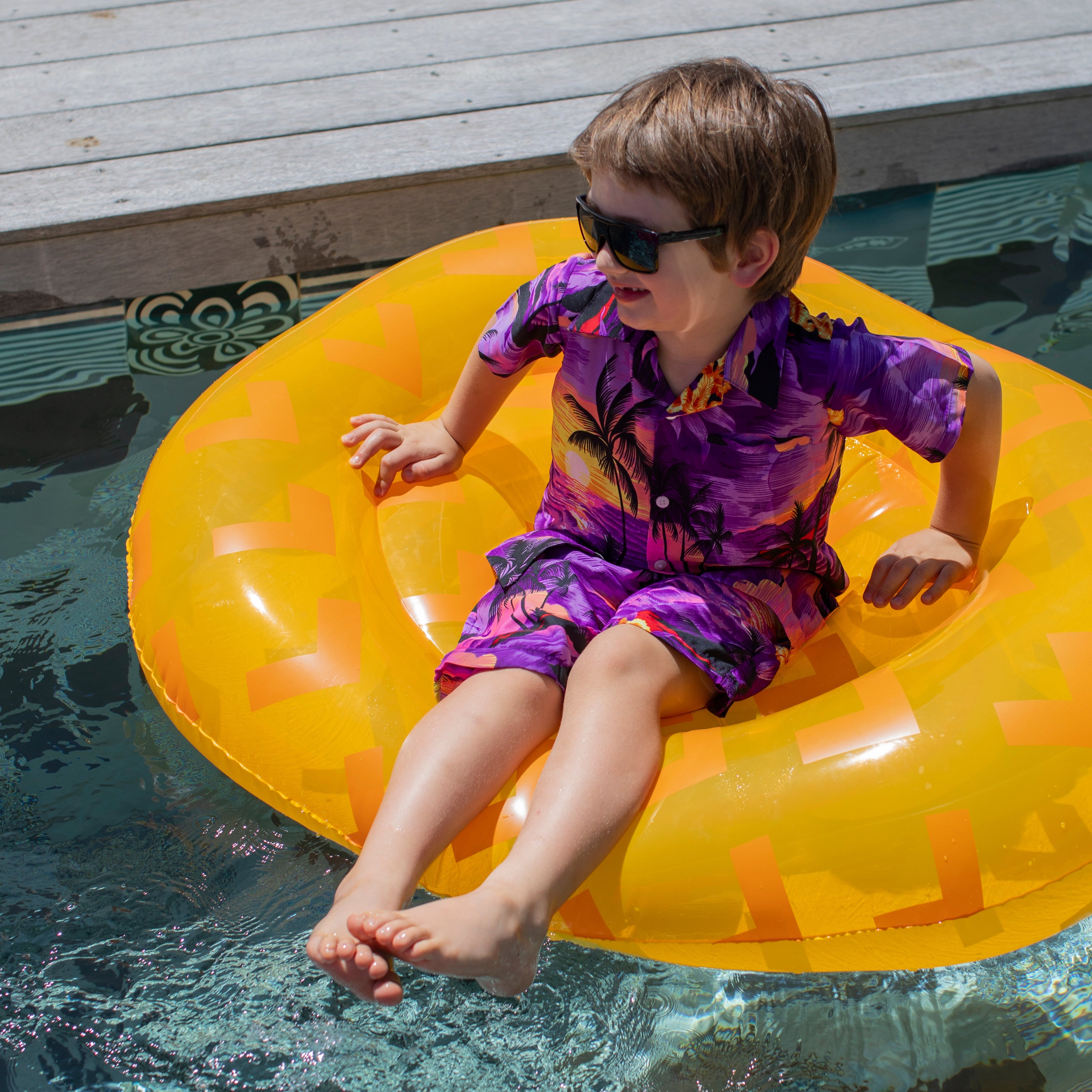 A young boy in a Sunset Purple Kids Shirt, featuring a bright design, sits on a yellow inflatable float in a pool, smiling as his feet dangle in the water beside a wooden deck.