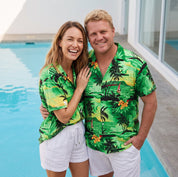 A smiling couple standing next to a swimming pool, wearing matching green tropical palm tree print short-sleeve button-up shirts and white shorts.