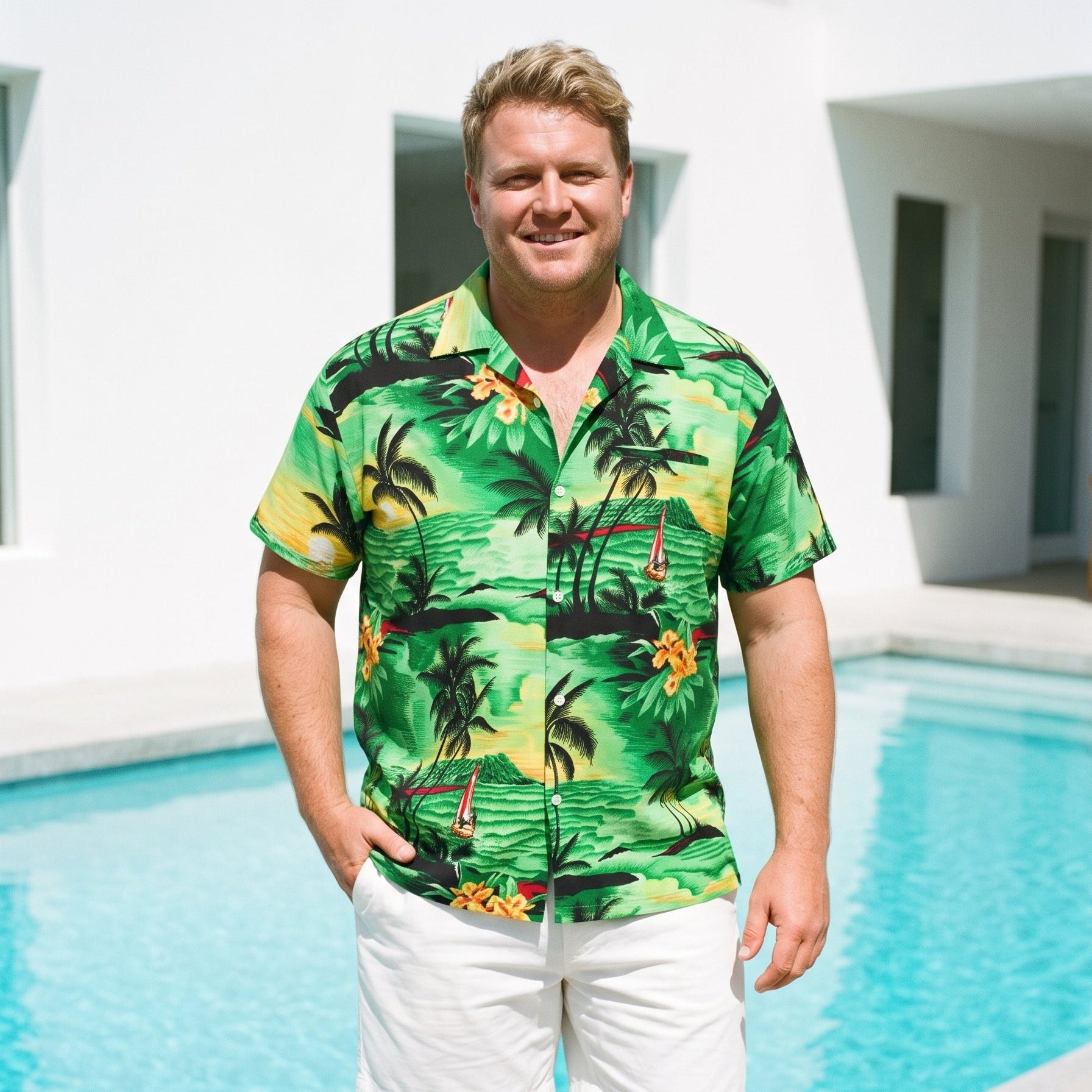 A man wearing a green tropical Hawaiian shirt with palm trees and yellow flowers, paired with white shorts, standing by a swimming pool in front of a modern white building.