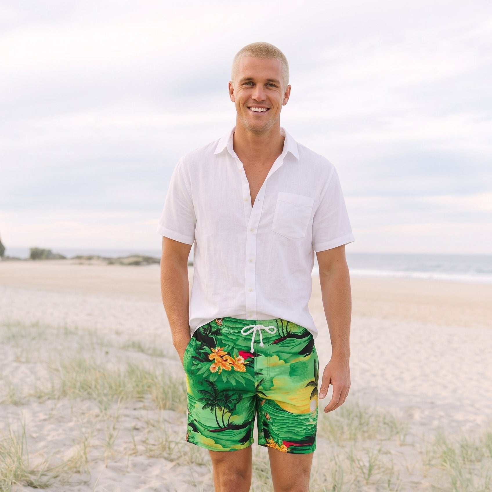 A man walking on a sandy beach wearing green tropical print swim trunks and a white short-sleeve button-down shirt.