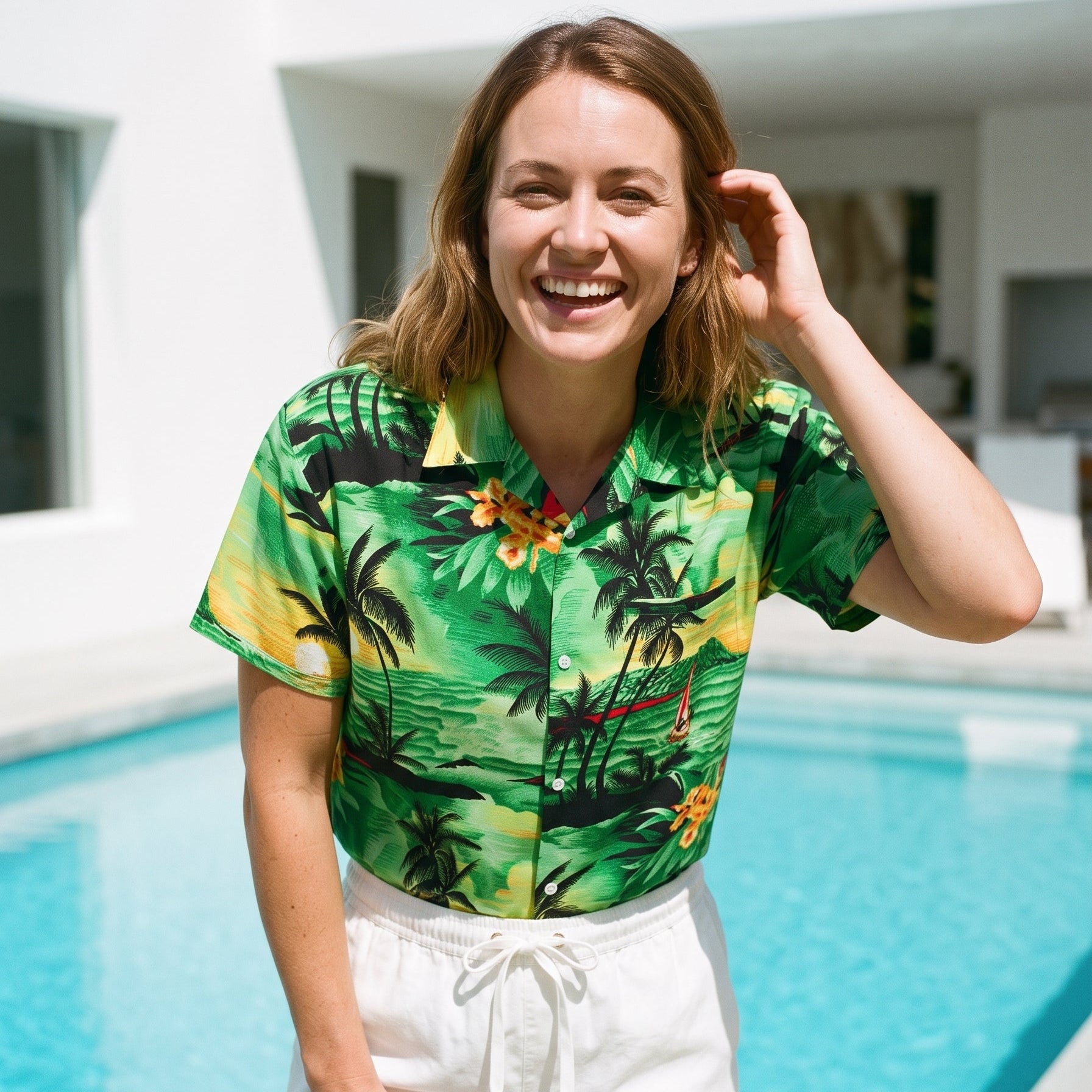 A woman smiling in front of a swimming pool, wearing a short-sleeve tropical print button-up shirt with white drawstring shorts.
