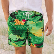 A man walking on a sandy beach wearing green tropical print swim trunks and a white short-sleeve button-down shirt.