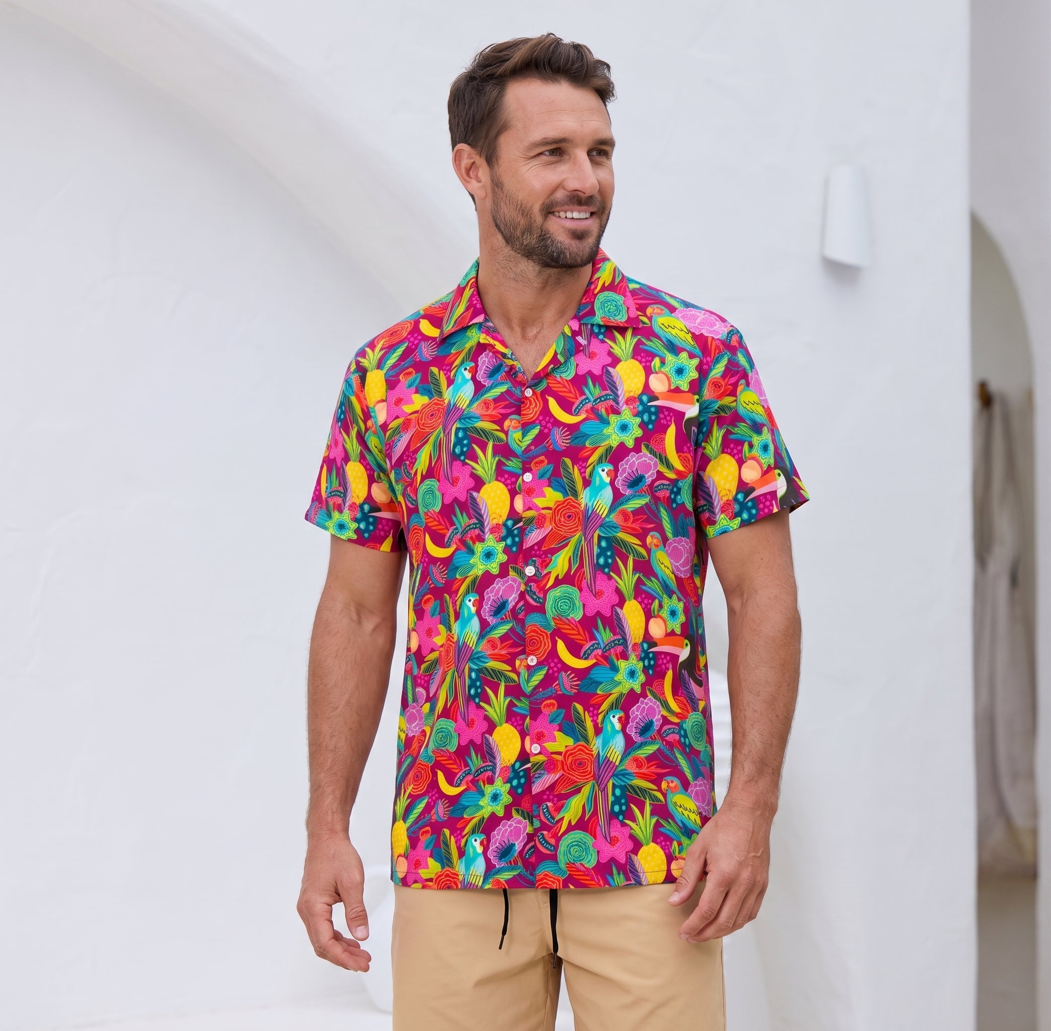 A man wearing a vibrant, multicolored tropical floral print short-sleeve button-down shirt and khaki pants, standing against a clean white stucco wall.