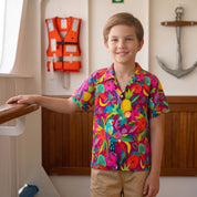 A young boy smiling and wearing a vibrant, multicolored tropical floral short-sleeve button-up shirt and khaki pants, standing indoors near a wooden railing with a nautical wall decor background.