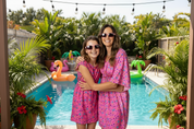 Two women in matching pink outfits with sunglasses against a light purple background