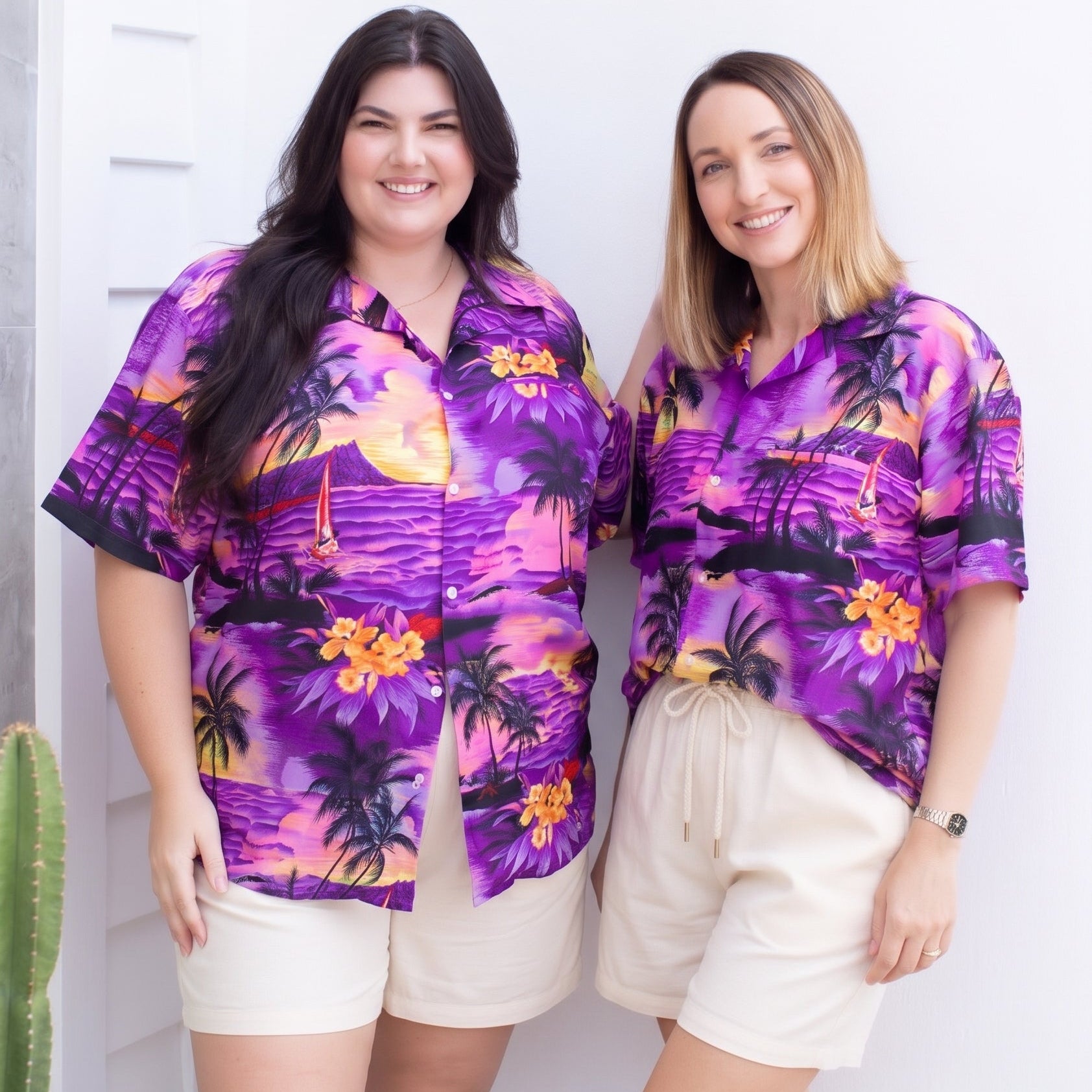 Two women standing side-by-side wearing matching purple tropical print short-sleeve button-up shirts and cream shorts, posing against a white wall.