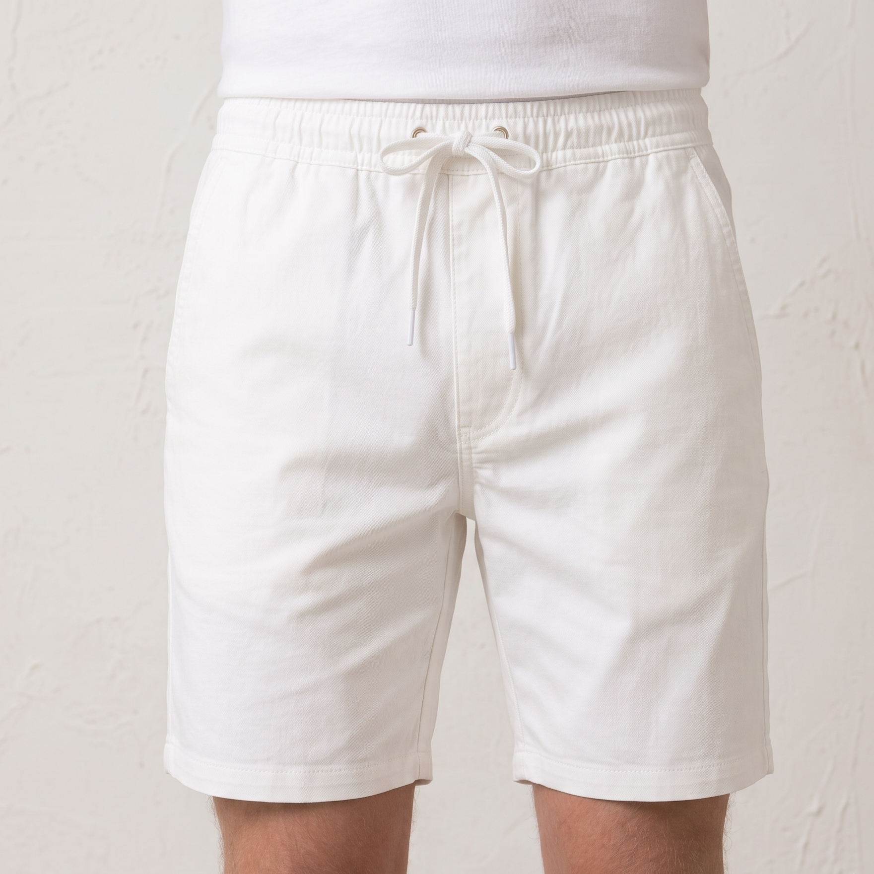 White drawstring shorts worn by a model, shown from the waist down against a textured off-white wall.