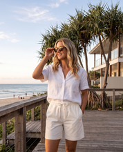 A woman stands on a wooden boardwalk by the beach wearing a white short-sleeve button-front shirt and white drawstring shorts. Palm trees, ocean, and a beach house are visible in the background.