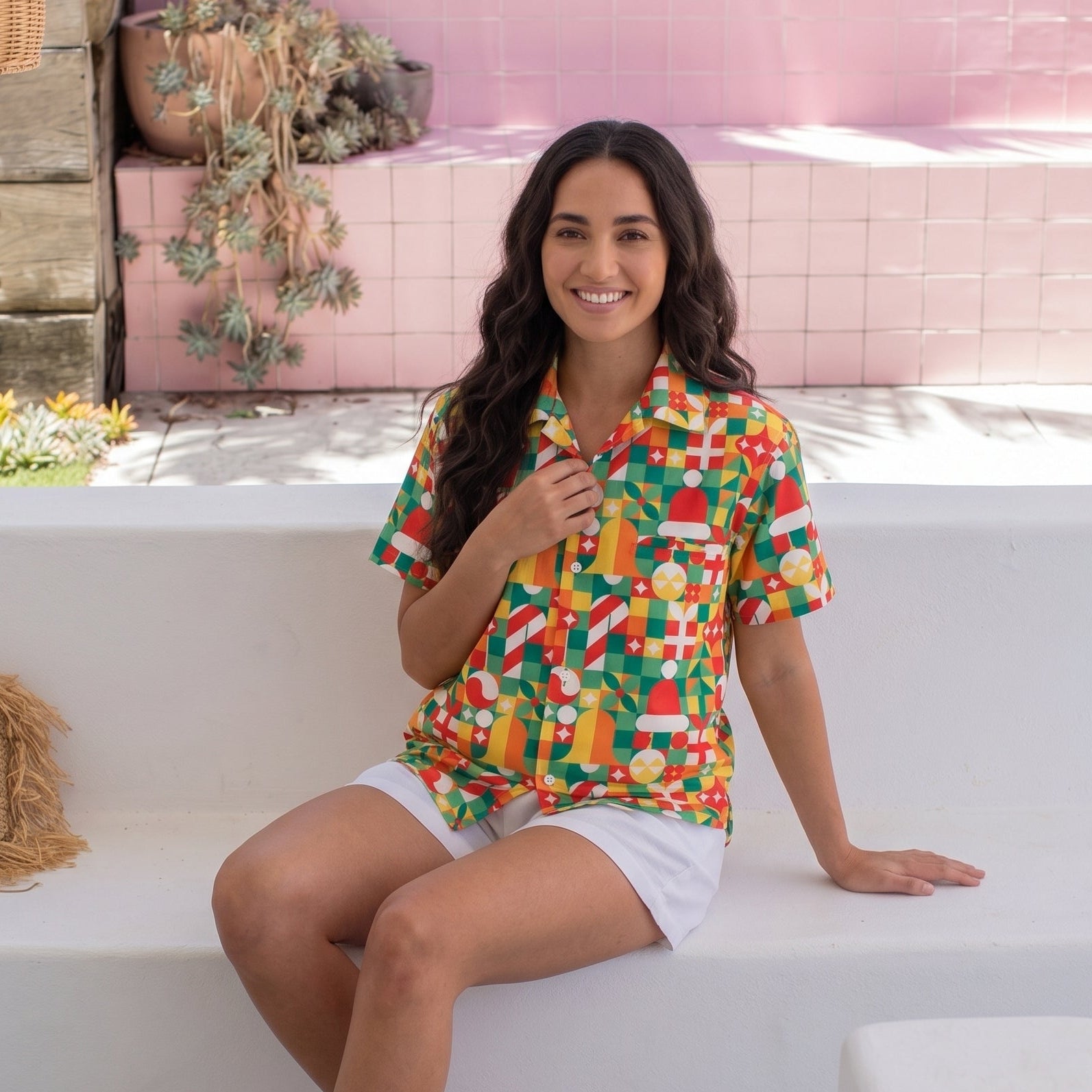 A woman sits on a white outdoor bench wearing a colorful short-sleeve button-up shirt with a bold tropical fruit and candy print, paired with white shorts and beige slide sandals, set against a pink tiled wall background.