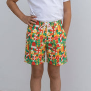 A young girl stands barefoot against a plain light gray wall, wearing a white short-sleeve t-shirt and colorful patterned shorts with a white drawstring.
