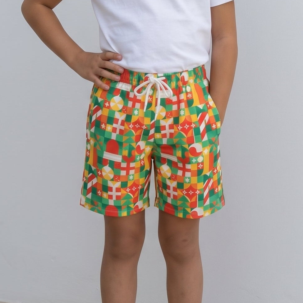 A young girl stands barefoot against a plain light gray wall, wearing a white short-sleeve t-shirt and colorful patterned shorts with a white drawstring.
